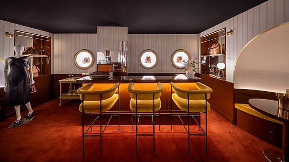 Luxurious room with yellow bar stools, mannequin in designer clothing, warm lighting, and handbags on display shelves.