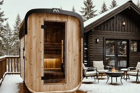 Outdoor sauna on a snowy wooden deck, next to a cabin with chairs around a fire pit, surrounded by snow-covered trees.