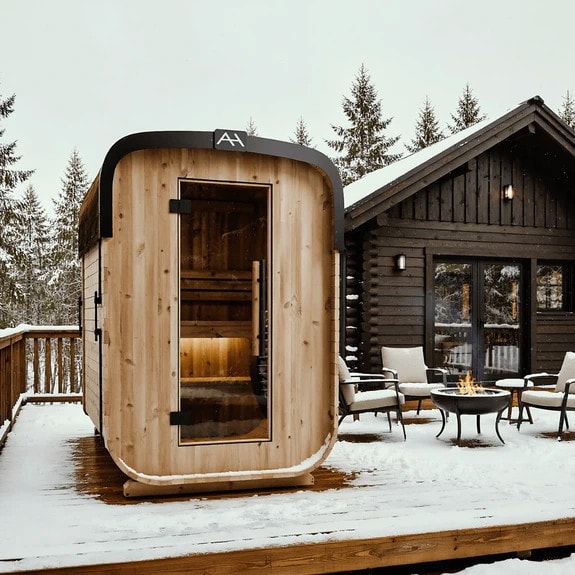 Outdoor sauna on a snowy wooden deck, next to a cabin with chairs around a fire pit, surrounded by snow-covered trees.