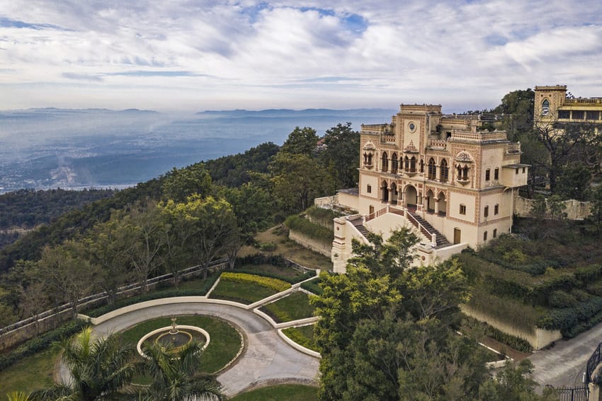Hilltop mansion surrounded by lush greenery and winding pathway with a distant view of the valley under a blue sky.