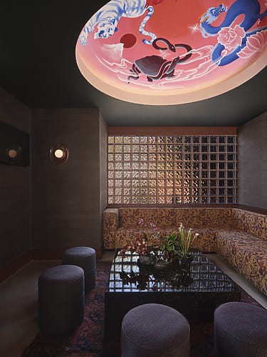 Cozy lounge with modern furniture, artistic ceiling mural, patterned seating, glass block wall, and soft lighting.