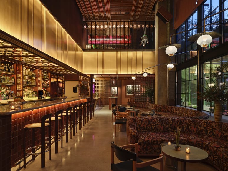 Modern bar interior with warm lighting, wooden barstools, and cozy patterned seating next to large windows at dusk