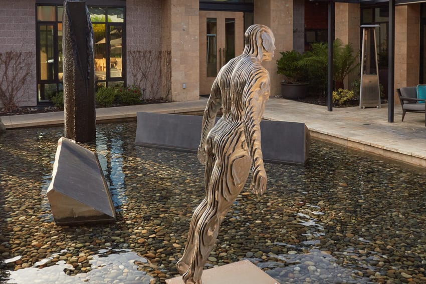 Modern sculpture of a man leaning forward in a shallow pebble-filled water feature, surrounded by contemporary architecture.