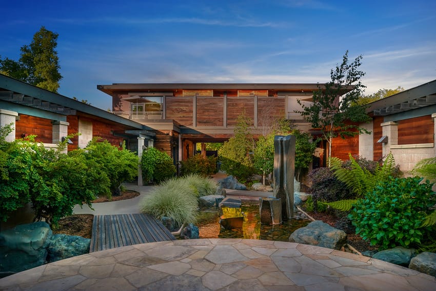 Modern building with rustic wood accents, lush gardens, a stone path, and a sleek water feature under a clear blue sky.