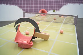 Abstract sculpture resembling a ping pong table with pink balls, a net, and unconventional shapes in an indoor setting.