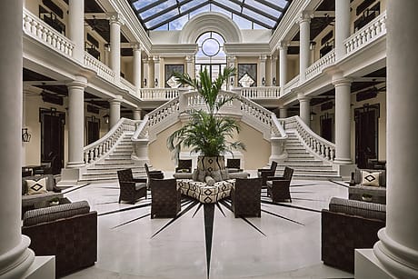 Elegant hotel lobby with grand staircases, tall columns, a central palm plant, and wicker seating under a glass roof.