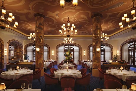 Elegant restaurant interior with ornate ceiling, chandeliers, and round tables adorned with white tablecloths and stylish chairs.