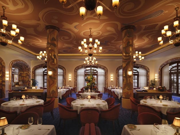 Elegant restaurant interior with ornate ceiling, chandeliers, and round tables adorned with white tablecloths and stylish chairs.