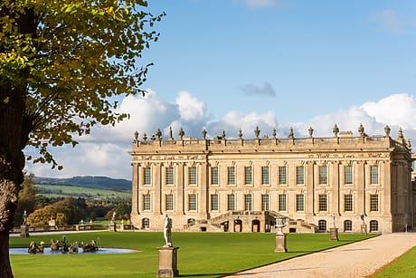 Grand historic estate with ornate architecture, manicured lawns, fountains, and sculptures under a clear blue sky.