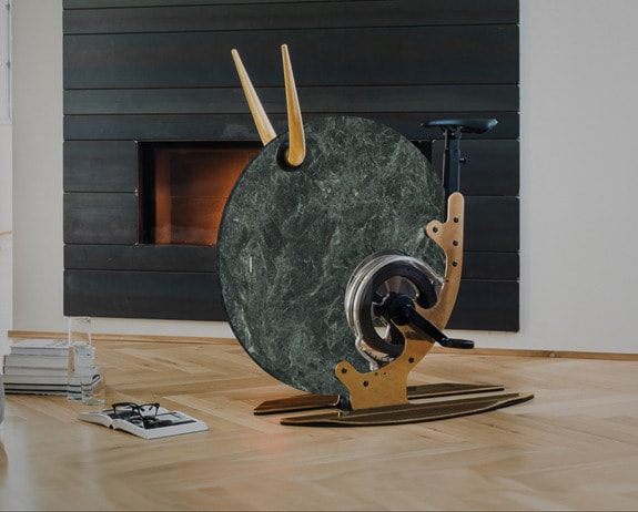 Luxury indoor exercise bike with a marble finish, placed on a wooden floor in front of a modern fireplace.
