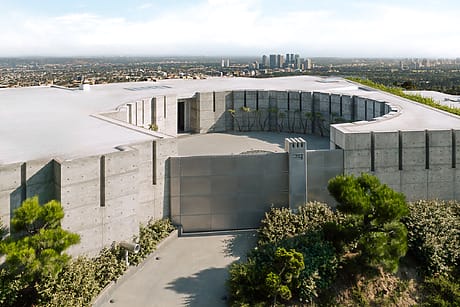 Modern concrete building with circular opening on top, surrounded by greenery, with city skyline in the background.