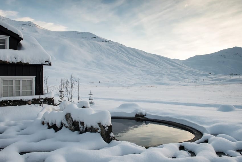 Snow-covered landscape with a cozy cabin, mountains, and a small round pond reflecting the serene winter scene.