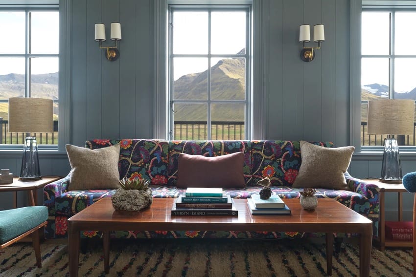 Colorful living room with floral sofa, wooden coffee table, and mountain view through three windows in the background.
