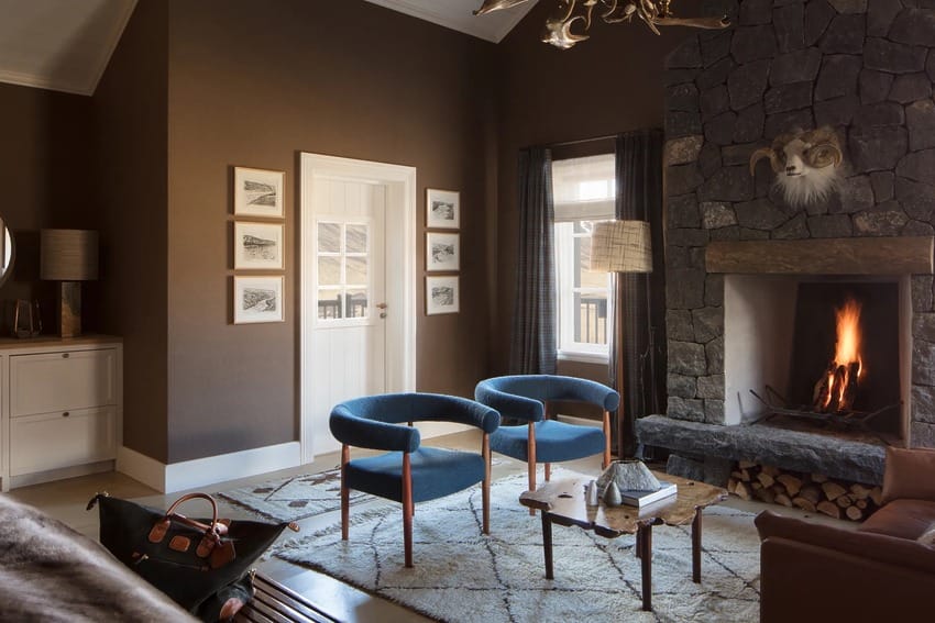 Cozy living room with stone fireplace, blue chairs, and framed pictures on brown walls. Logs stored below the mantel.