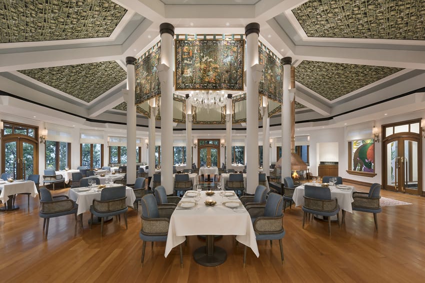 Luxurious restaurant dining room with elegant decor, round tables, blue chairs, large chandelier, and intricate ceiling patterns