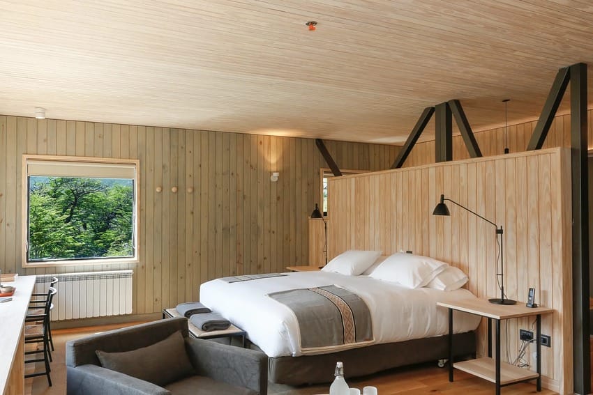 Modern hotel room with wooden interior, large bed, couch, desk, and forest view through window.