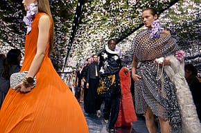 Models in vibrant, floral-inspired outfits walk a runway under a canopy of flowers during a fashion show.