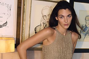 Woman in a beige dress posing indoors with framed artwork in the background.