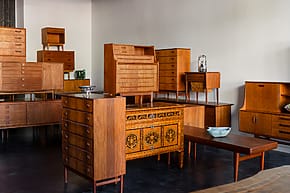 Room filled with vintage wooden furniture including cabinets, drawers, and tables arranged in a minimalist interior setting.