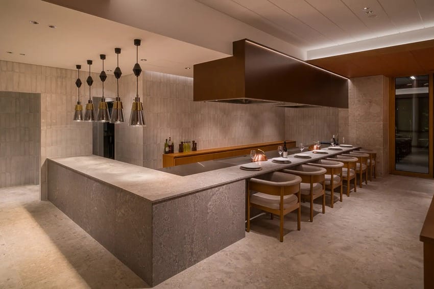 Modern restaurant interior with a long counter, bar stools, pendant lights, and neutral-toned decor.