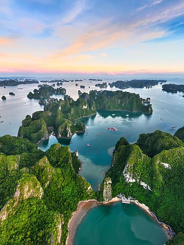 Aerial view of lush green islands and turquoise water under a colorful sunset sky in Ha Long Bay, Vietnam.