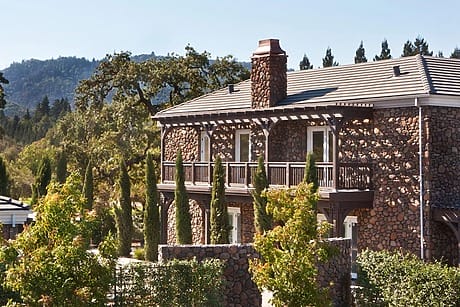 Stone house with chimney surrounded by greenery and trees against a backdrop of hills and clear sky.
