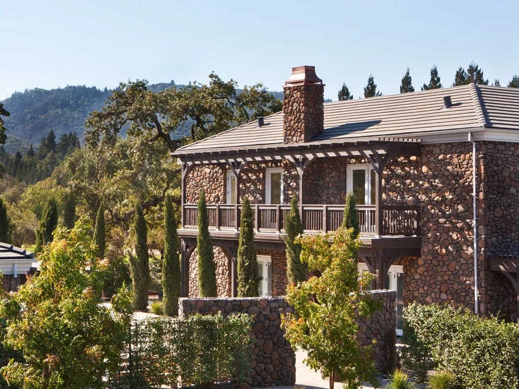Stone house with chimney surrounded by greenery and trees against a backdrop of hills and clear sky.