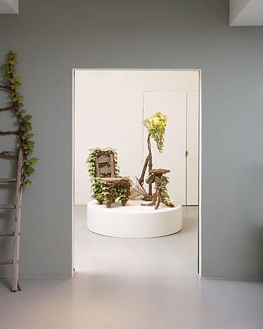 Art installation with wooden structures and green vines on a circular pedestal in a minimalist, gray-walled room.