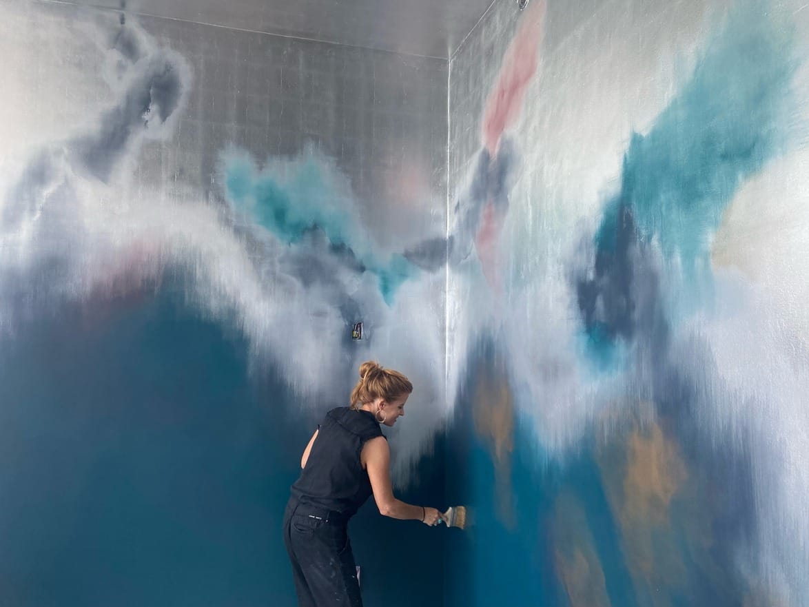 Artist painting a vibrant abstract mural with blue, white, pink, and gold hues on a large wall in an indoor space.