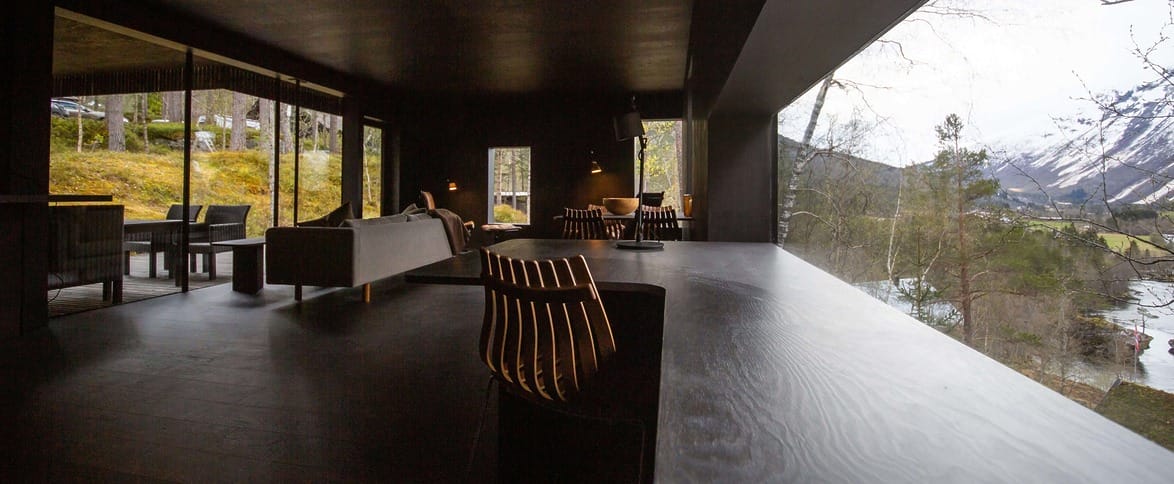 Modern living room with dark interior, large windows showing scenic mountain and forest views.