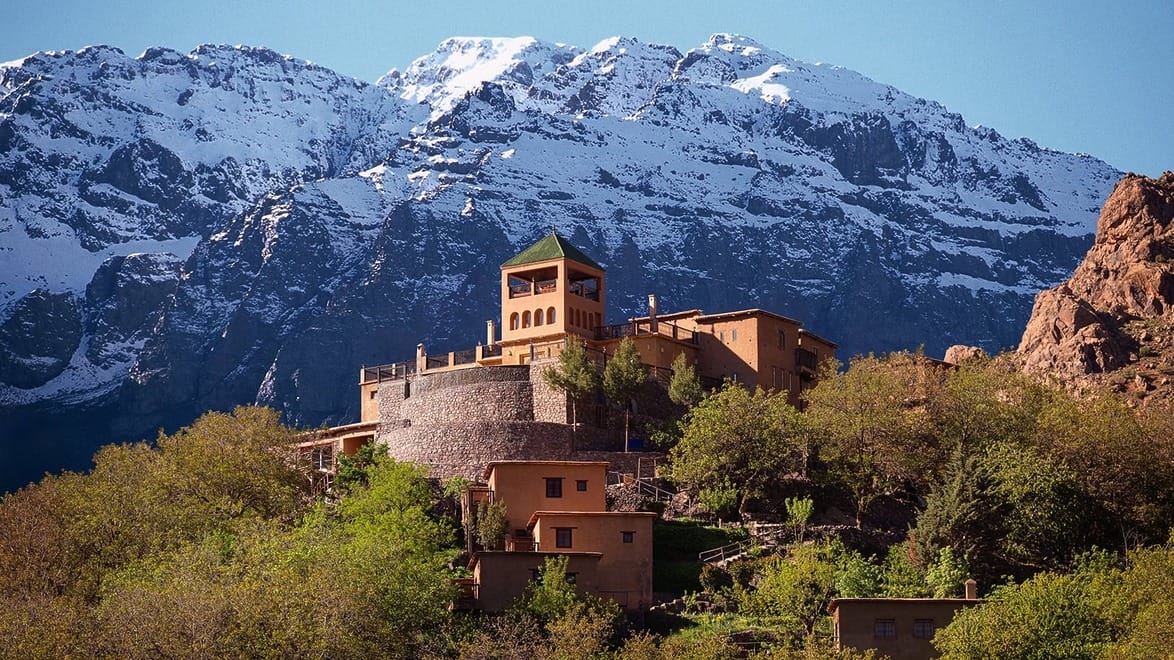 Mountainous landscape with a large, castle-like building surrounded by trees, set against a backdrop of snow-capped peaks.