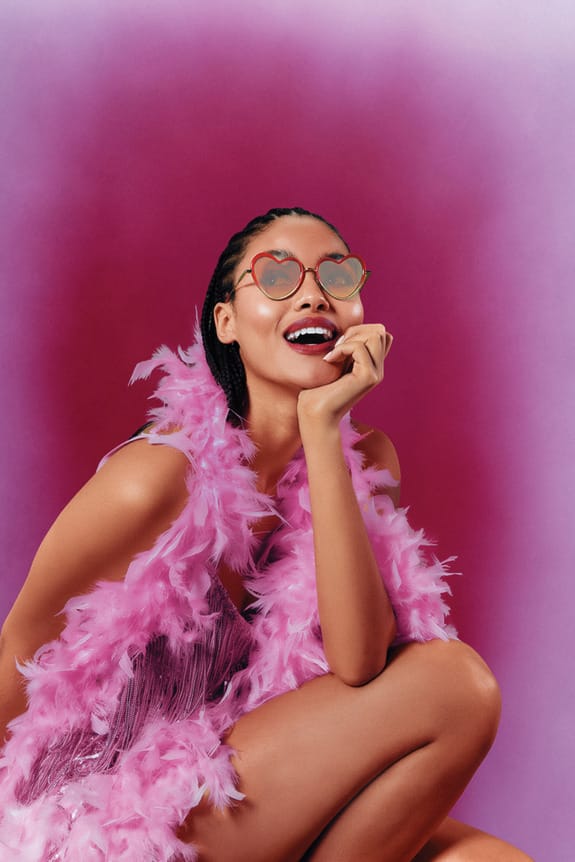 Woman in heart-shaped sunglasses and pink boa, smiling against a pink gradient background.