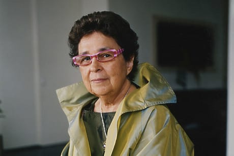 Person wearing a yellow jacket and pink glasses with short dark hair looking at the camera in an indoor setting