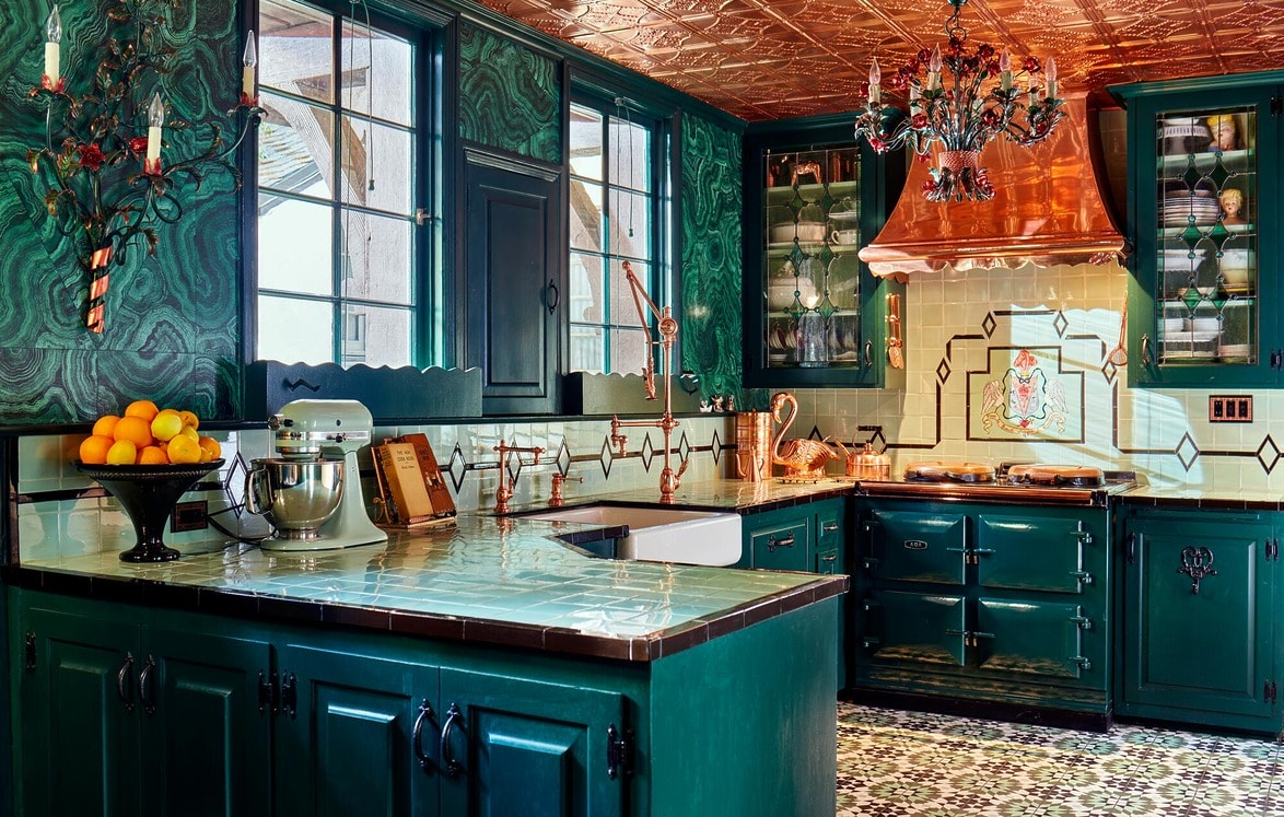 Eclectic kitchen with emerald cabinets, copper range hood, patterned floor tile, and a decorative chandelier.