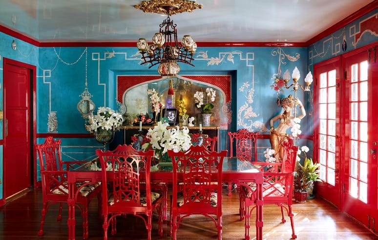 Vibrant dining room with red chairs, glass table, blue walls, floral arrangements, and ornate chandelier.