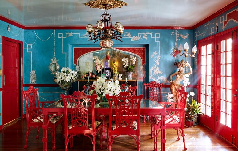 Vibrant dining room with red chairs, glass table, blue walls, floral arrangements, and ornate chandelier.