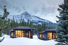 Modern cabins with large windows in snowy forest, mountain backdrop, twilight sky.