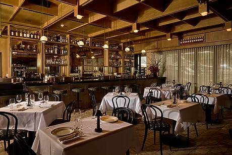 Cozy upscale restaurant interior with elegantly set tables, ambient lighting, and a fully stocked bar in the background.