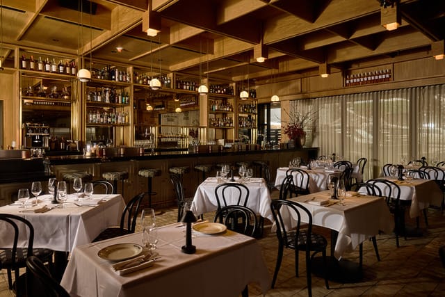 Cozy upscale restaurant interior with elegantly set tables, ambient lighting, and a fully stocked bar in the background.
