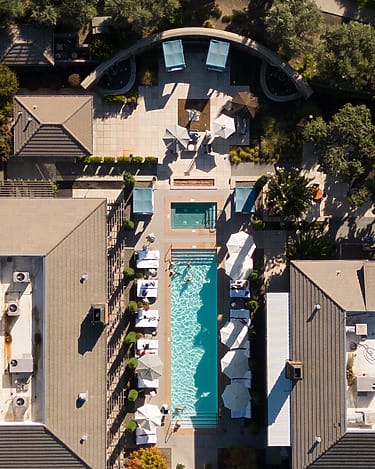 Aerial view of a modern pool area with loungers, surrounded by buildings and trees on a sunny day.