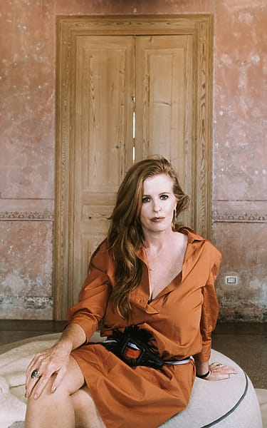 Woman in orange dress sitting in a rustic room with pottery on wooden tables in the background.