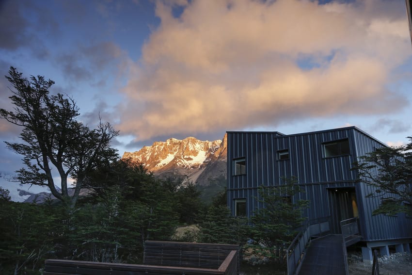 Modern building with a scenic view of mountains and a vibrant sky at sunset, surrounded by lush trees in the foreground.