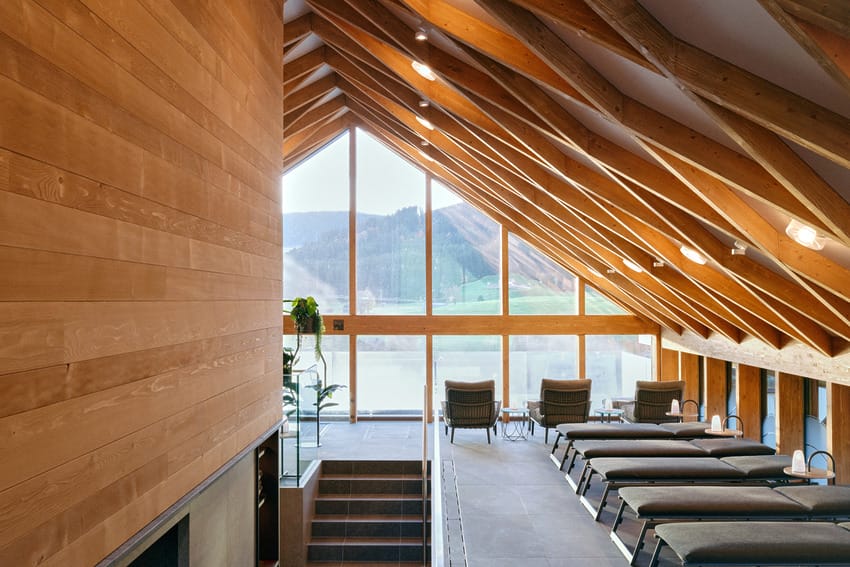 Modern spa relaxation room with large windows, wooden beams, and lounge chairs overlooking scenic outdoor view.