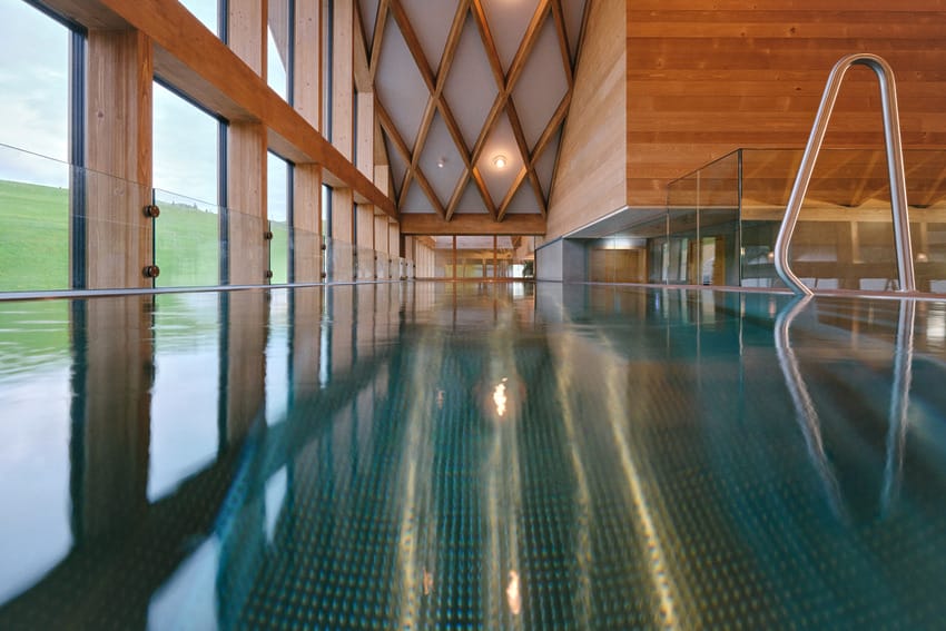 Indoor pool with wooden walls and large windows offering a view of the countryside, reflections on the water's surface.