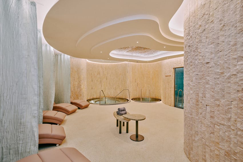 Luxury spa room with reclining chairs, stone walls, and hot tubs under a stylish, modern ceiling design.