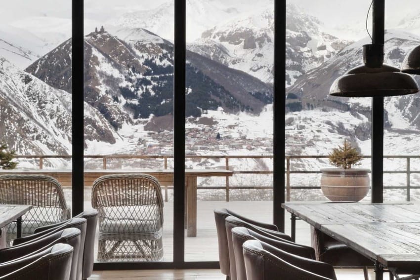 Mountain view from large windows in a cozy restaurant with wooden tables and wicker chairs, snowy peaks in the background.