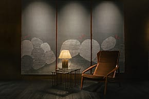 Cozy room with a wooden chair, side table, and lamp in front of a textured wall with abstract designs.