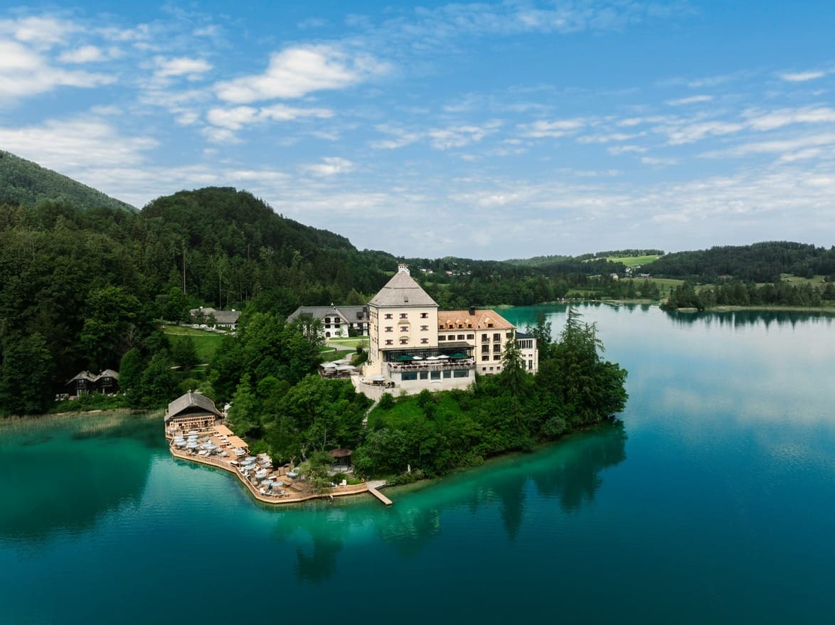 Aerial view of a lakeside chateau with lush green surroundings and clear blue water under a partly cloudy sky.