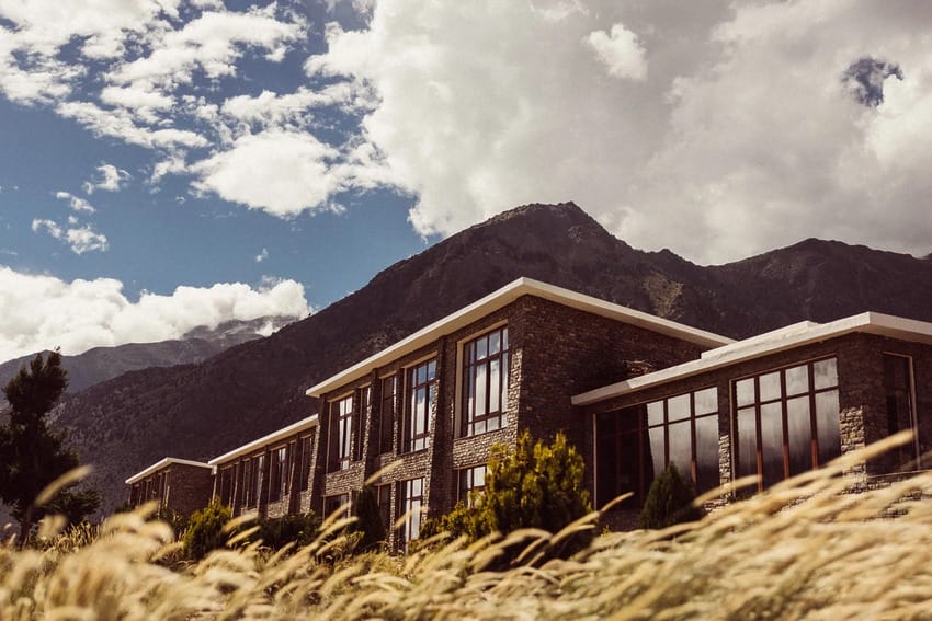 Modern building with large windows in front of a mountain under a cloudy sky, surrounded by tall grass.