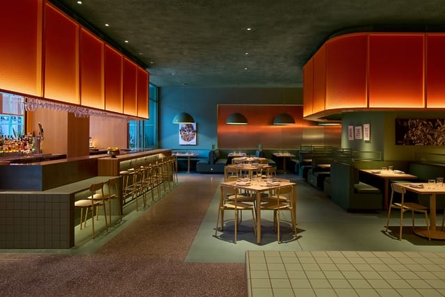 Modern restaurant interior with wooden tables, chairs, and a bar, featuring orange lighting and green accents.